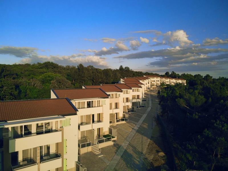 Row of modern holiday apartments with red roofs in a green, wooded landscape under a clear sky.