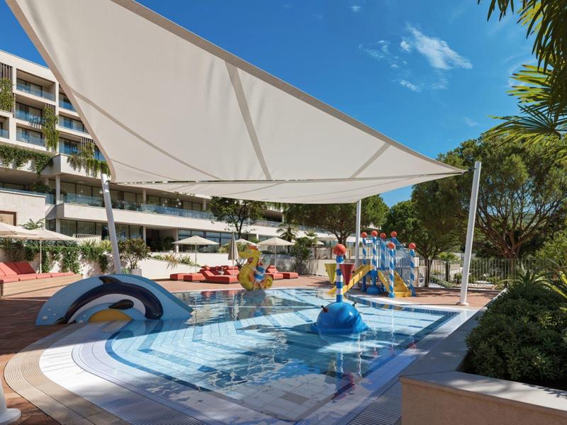 Hotelový bazén s vodním hřištěm a sluneční plachtou před vícepodlažní budovou.