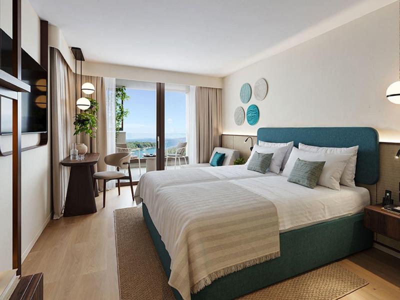 Moderní hotelový pokoj s velkou manželskou postelí, psacím stolem a balkonem s výhledem na moře.