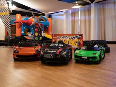 Drei kleine Elektrowagen in Orange, Schwarz und Grün stehen auf Holzfußboden vor einer Hüpfburg.