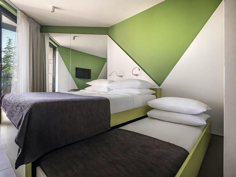 Modernes Schlafzimmer mit Doppelbett, zusätzlichem Einzelbett, grünen Akzenten und Balkonblick.