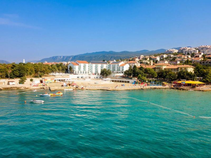 Klares Meer mit Küste, Hotels und Häusern unter blauem Himmel und Bergen im Hintergrund.