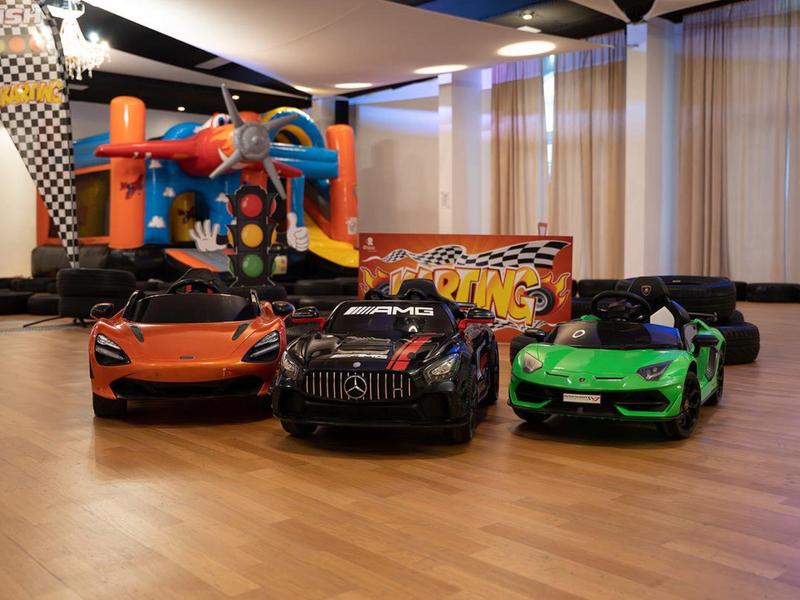 Drei kleine Elektrowagen in Orange, Schwarz und Grün stehen auf Holzfußboden vor einer Hüpfburg.