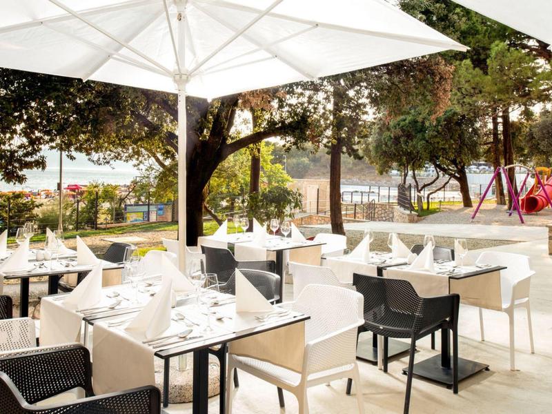 Freiluftrestaurant mit weißen Tischen, Stühlen, Sonnenschirm und Bäumen, Hintergrund mit Spielplatz.