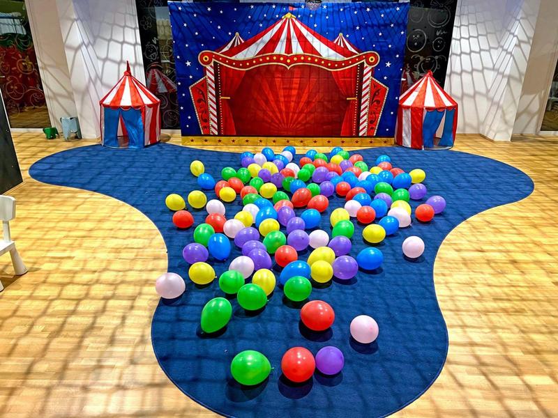 Bunte Luftballons auf blauem Teppich vor Zirkuszelt-Kulisse mit roten und weißen Zelten.