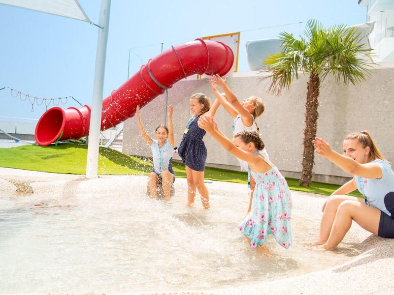 Kinder spielen im flachen Wasserbecken neben einer roten Rutsche und einer Palme.