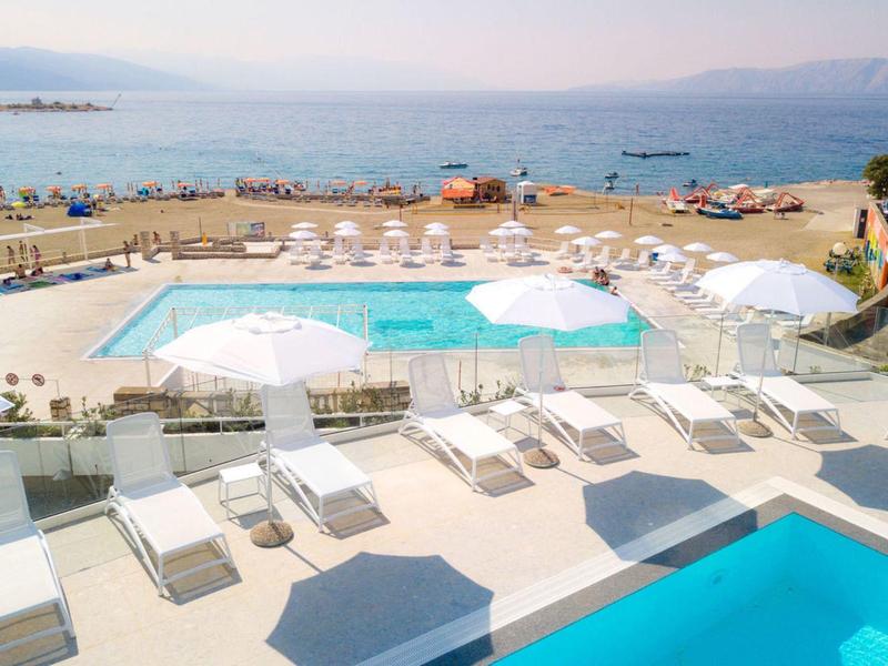Poolbereich mit weißen Liegestühlen und Sonnenschirmen, Meerblick und klarem Himmel im Hintergrund.