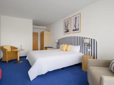 Pokój hotelowy z podwójnym łóżkiem, niebieskim dywanem i nowoczesnym wyposażeniem w neutralnych kolorach.
