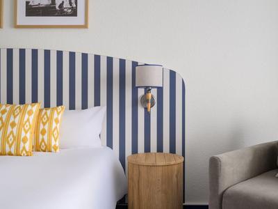 Pokój hotelowy z pasiastym zagłówkiem, białą pościelą, żółtymi poduszkami wzorzystymi i szarym fotelem.
