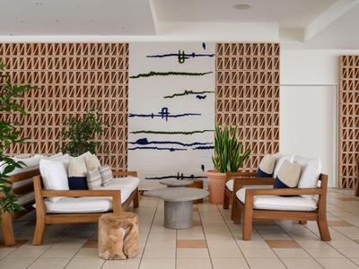 Elegancka lobby hotelowa z drewnianymi meblami, białymi poduszkami, roślinami doniczkowymi i abstrakcyjną sztuką ścienną.