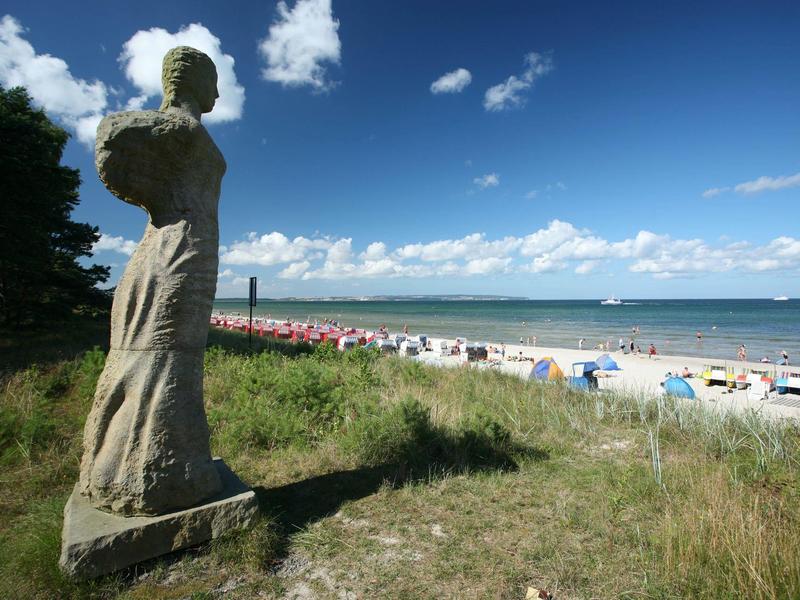 Eine steinerne Statue steht auf einem bewachsenen Hügel mit Blick auf einen Strand und das Meer.