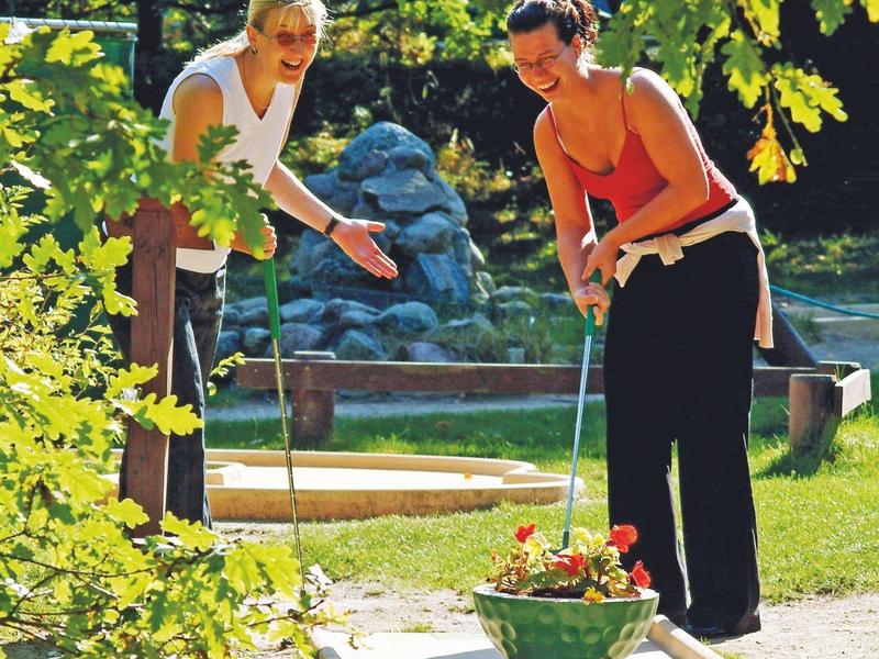 Zwei Frauen spielen Minigolf auf einem sonnigen Platz mit grüner Umgebung und Bäumen.