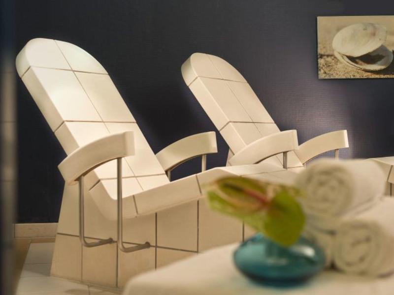 Zwei weiße Sesseln mit Armlehnen vor dunkler Wand, vorne grüne Vase mit Blume und weiße Handtücher.