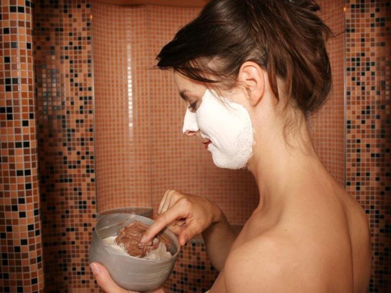 Frau trägt Gesichtsmaske auf, hält Schale mit cremiger Substanz, braune Mosaikfliesen im Hintergrund.