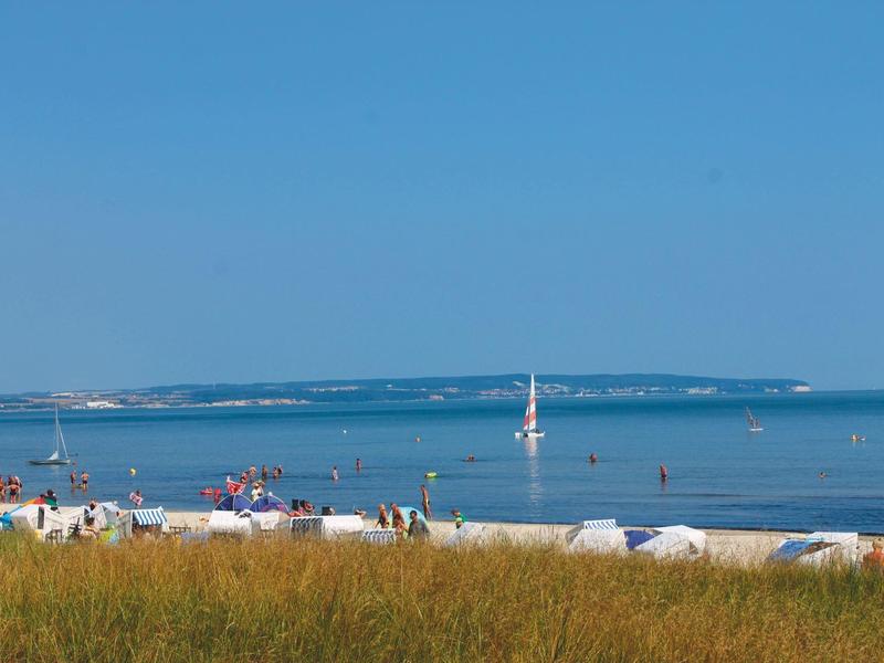 Strand mit weißen Sonnenschirmen, gelbem Gras im Vordergrund und blauem Meer mit Segelbooten im Hintergrund.