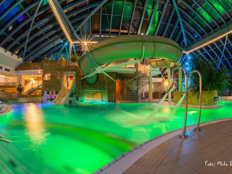 Innenhallenwasserpark mit grün beleuchtetem Pool und Rutschen unter einer gläsernen Kuppel.