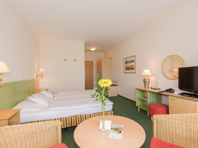 Hell eingerichtetes Hotelzimmer mit Doppelbett, Tisch, Stühlen, TV und gelben Blumen auf dem Tisch.