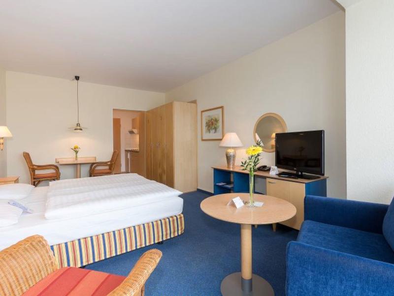 Helles Hotelzimmer mit Doppelbett, blauem Teppich, runder Tisch, blauer Couch und TV auf Kommode.