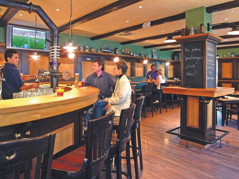 Gemütliche Bar mit Holzmöbeln, Tresen und Gästen, warmes Licht und Sport im Hintergrund.