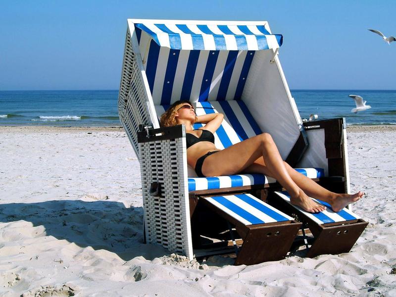 Frau in Bikini entspannt in blau-weiß gestreiftem Strandkorb am Sandstrand mit Meer im Hintergrund.