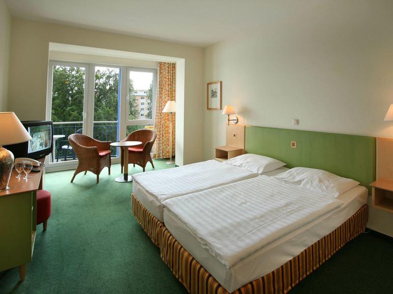 Helles Hotelzimmer mit Doppelbett, grünem Teppich, zwei Stühlen vor Fenster und sanfter Beleuchtung.