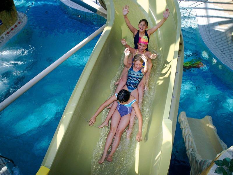 Vier Personen rutschen hintereinander auf einer gelben Wasserrutsche im Schwimmbad.