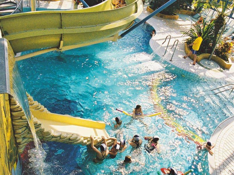 Innen schwimmbad mit großer gelber Wasserrutsche und mehreren schwimmenden Personen im Becken.