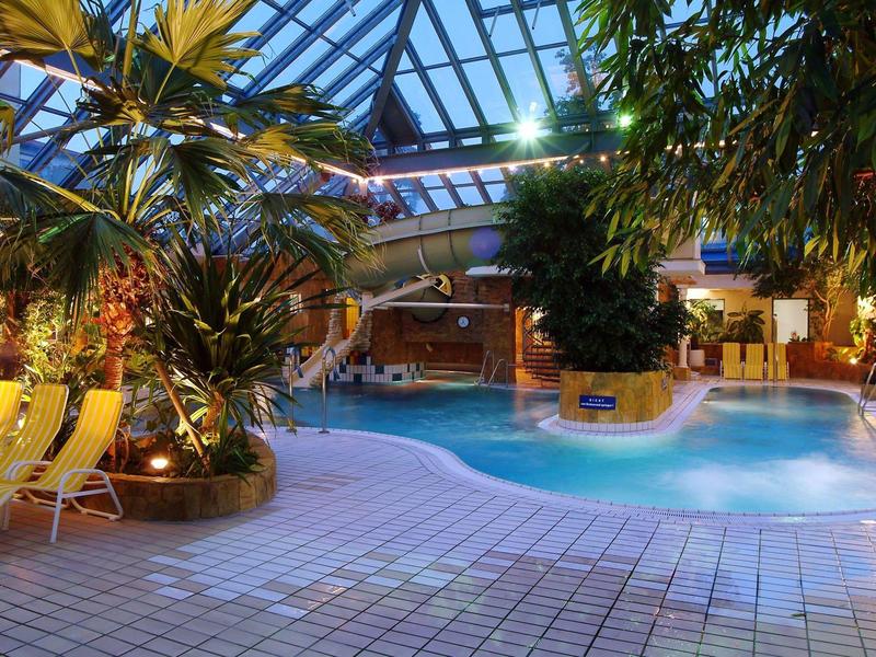 Innenraum eines tropischen Hallenbades mit Pool, Pflanzen und gelben Liegestühlen unter Glasdach.