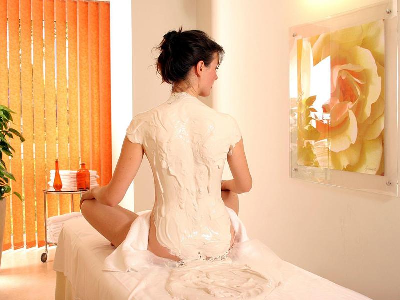 Frau mit weißem Kleid sitzt auf Massageliege in heller, gemütlicher Wellnessraumatmosphäre.