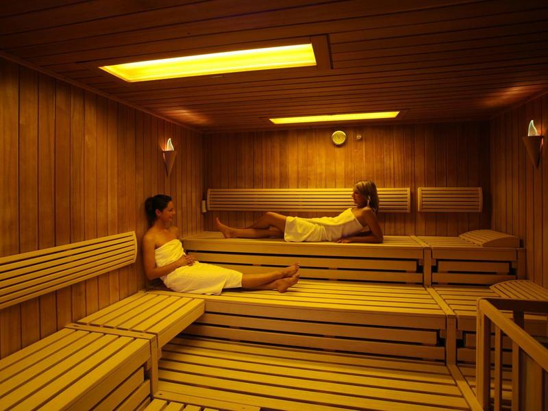 Saunaraum mit Holzverkleidung, zwei Frauen sitzen auf mehrstufigen Holzbänken, warmes Licht.