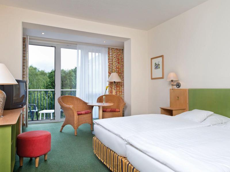 Helles Hotelzimmer mit Doppelbett, grünem Teppich, zwei Stühlen, Fernseher und Balkon mit Waldsicht.