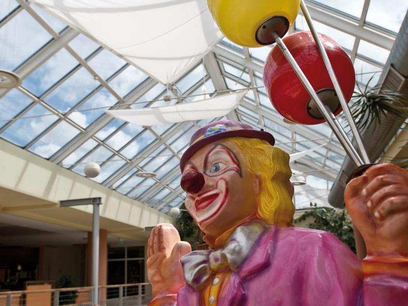Bunter Clown mit Luftballons in Einkaufszentrum unter Glasdach, Kinder spielen im Hintergrund.