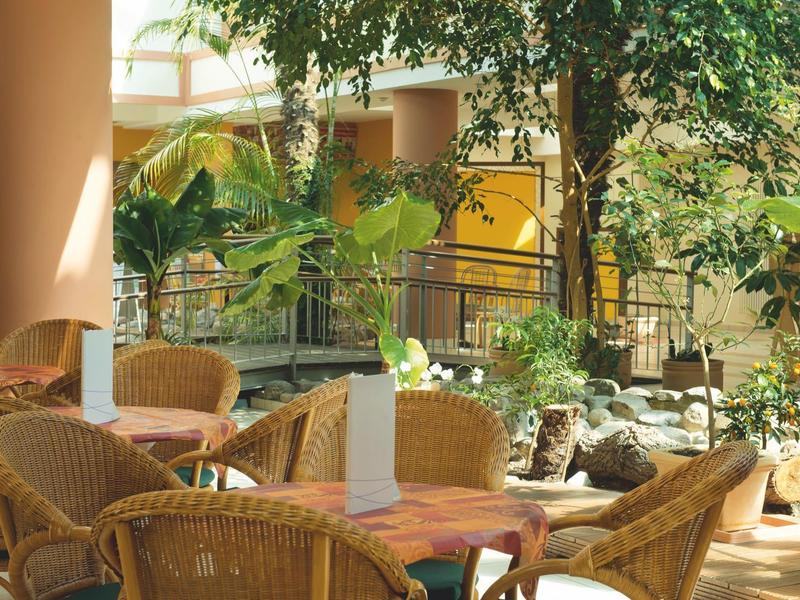 Gemütliche Sitzgruppe mit Korbstühlen und Tischen umgeben von üppigen grünen Pflanzen in hellem Raum.