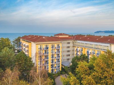 Hotelgebäude mit roten Dächern inmitten grüner Bäume und Blick aufs Meer im Hintergrund.