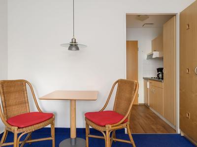 Small dining area with two chairs with red cushions, wooden table, and kitchenette in the background.