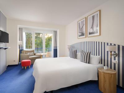 Modern hotel room with double bed, blue carpet, and large window overlooking greenery.