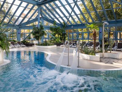 Indoor pool with waterfalls and lounge chairs under a glass roof structure.