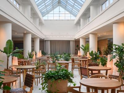 Bright indoor courtyard with wooden tables, chairs, and many green plants under a glass roof.