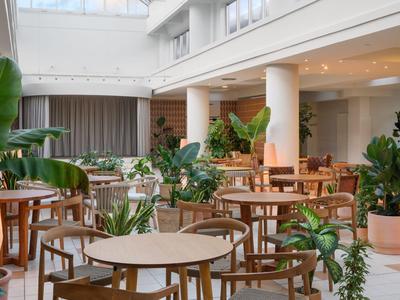 Bright hotel dining area with wooden tables, chairs, and numerous green plants.