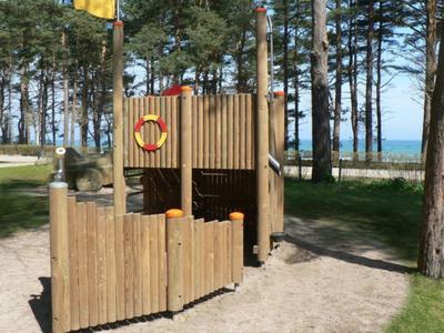 Ein hölzernes Kinderspielschiff in einem sandigen Bereich mit Bäumen und Meer im Hintergrund.