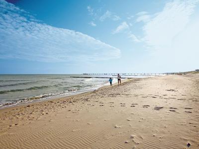 Weite Sandstrand mit Fußspuren, ruhigem Wasser und zwei Personen am Horizont.