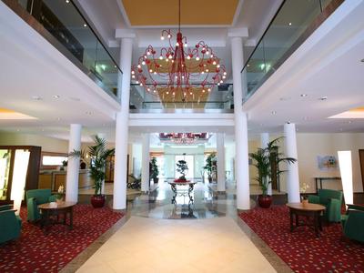 Heller Hotellobby mit roten Teppichen, grünen Sesseln und einer großen Kronleuchterdecke.