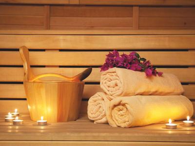 Holzsauna mit drei gerollten Handtüchern, Eimer, Schöpfkelle und dekorativen Blumen, beleuchtet von Teelichtern.