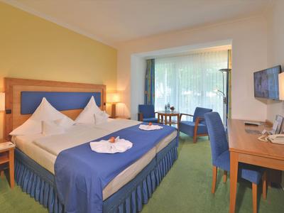 Helles Hotelzimmer mit Doppelbett, blauen Möbeln und Blick auf einen Balkon mit Garten.