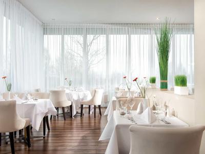 Helles Restaurant mit weißen Stühlen, Tischdecken, Holzfußboden und großen Fenstern mit Vorhängen.