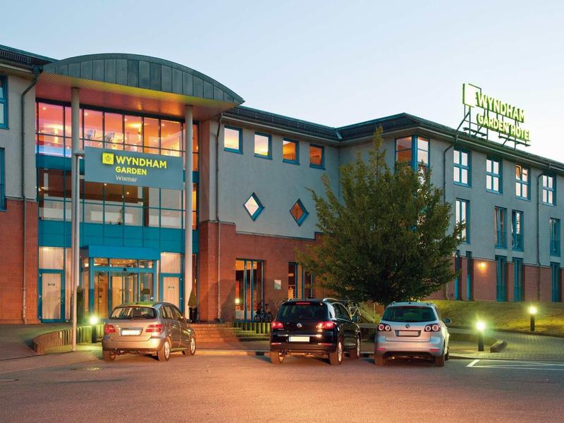 Modernes, mehrstöckiges Hotelgebäude mit beleuchtetem Eingang und geparkten Autos davor bei Dämmerung.