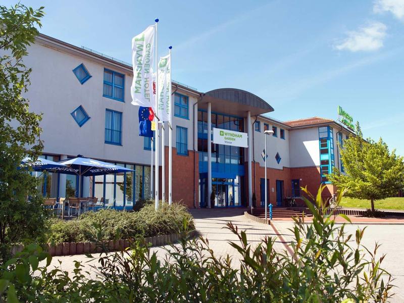 Modernes Gebäude mit blau-rotem Eingang, Fahnenmasten und grüner Umgebung unter blauem Himmel.