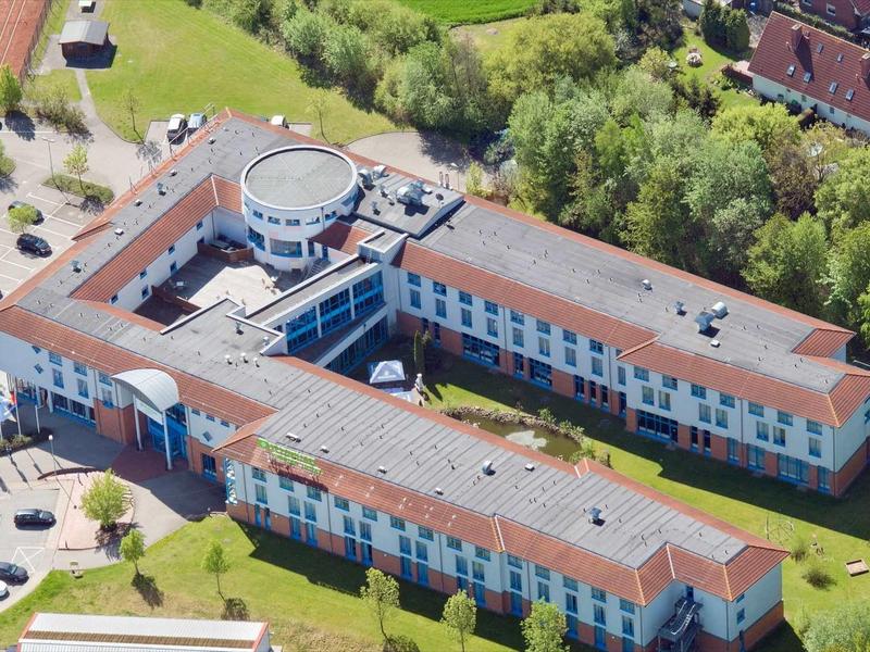 Großes, U-förmiges Gebäude mit rotem Dach, umgeben von Grünflächen und Parkplätzen.