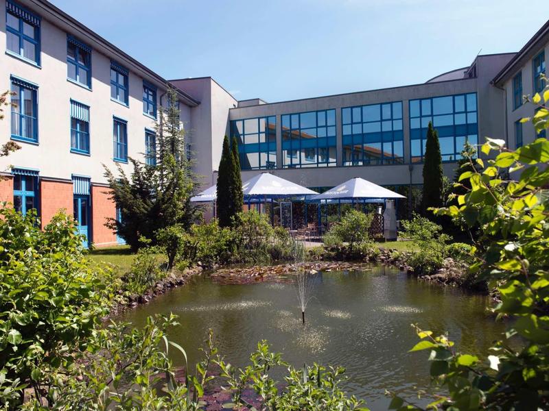 Modernes Gebäude mit Glasfassade, umgeben von Garten und Teich mit grüner Vegetation.