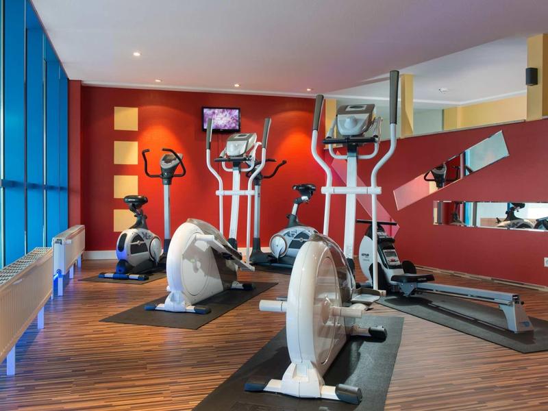 Fitnessraum mit roten Wänden, großen Fenstern und verschiedenen Trainingsgeräten auf Holzfußboden.